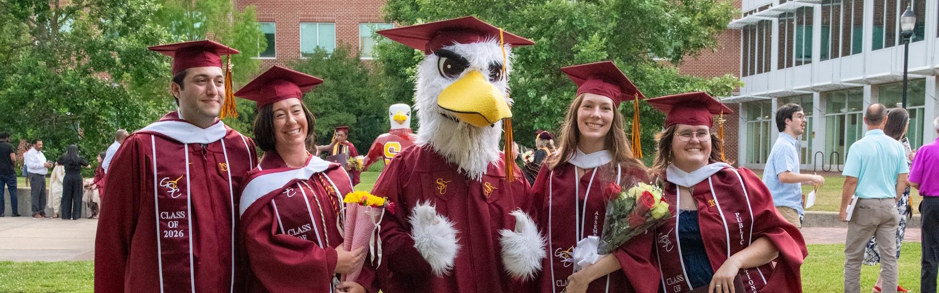 4 students in a graduation atire pose for photo with Sammy the Sea Gull