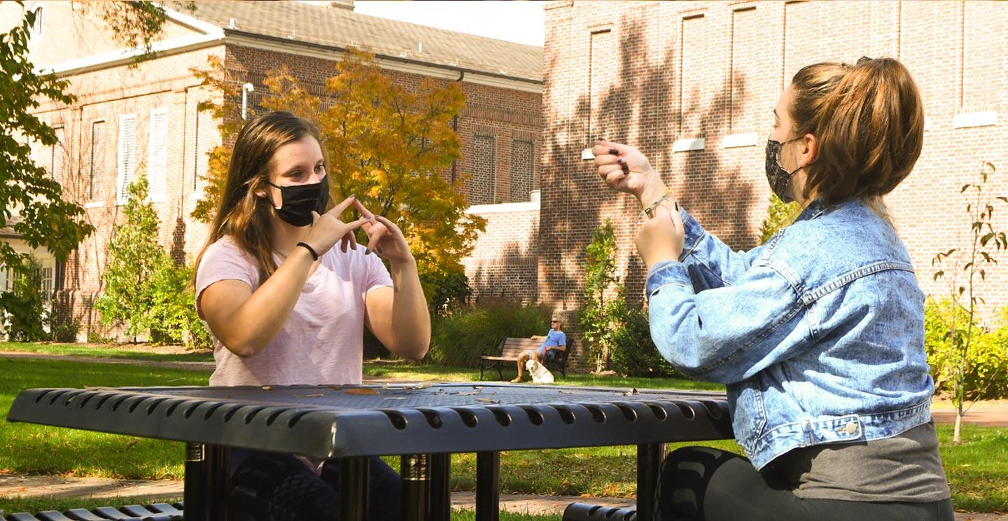 Two students at outside table use sign language
