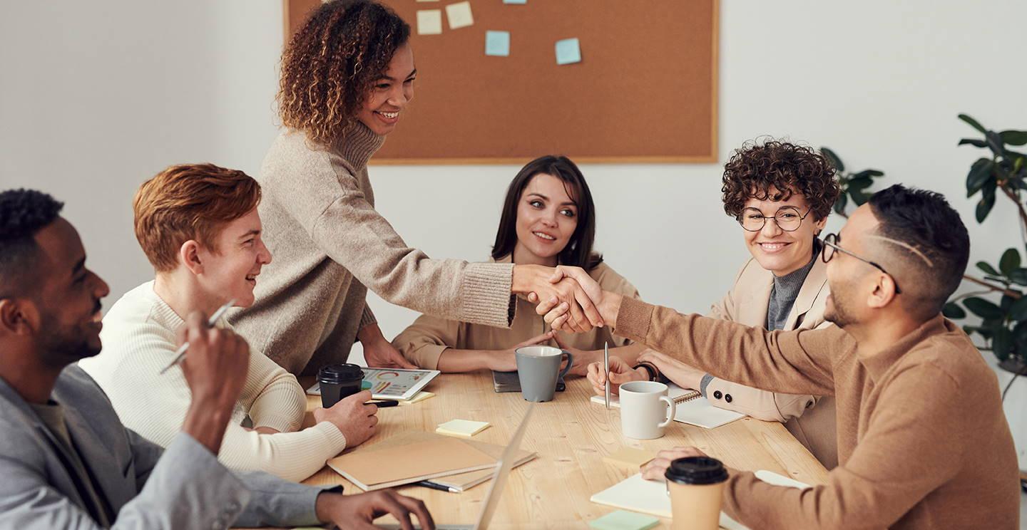 Group of people sit around table as one person standing is shaking the hand of someone sitting